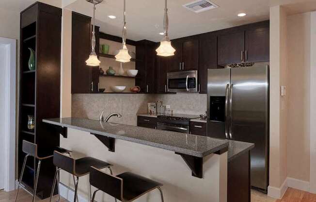 A modern kitchen with dark cabinets, stainless steel appliances, a breakfast bar with pendant lights, and built-in open shelves.