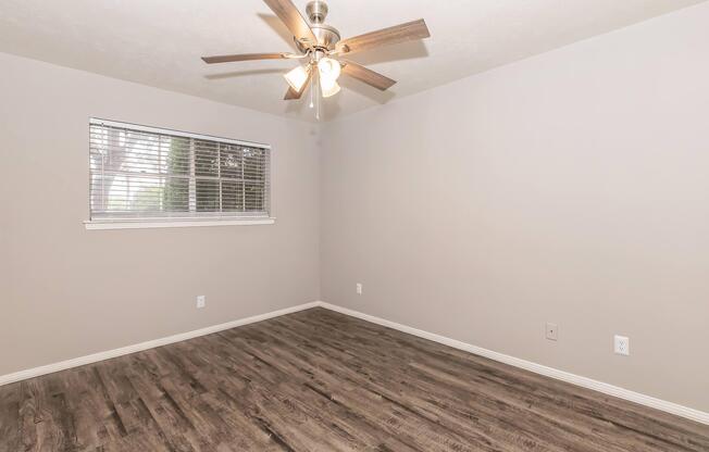 A light and airy empty room featuring a ceiling fan with wooden blades, a window with blinds allowing natural light, and light gray walls. The floor is covered with laminate wood-style flooring, creating a warm and inviting atmosphere.