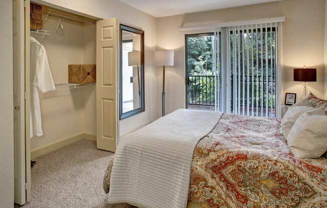 A cozy bedroom here at Heritage Hills showcasing a patterned bedspread with layered pillows, soft carpeted flooring, and neutral walls. The room features an open closet with shelving, a full-length wall mirror, bedside lighting, and sliding glass doors with vertical blinds that open to a private balcony overlooking mature trees.