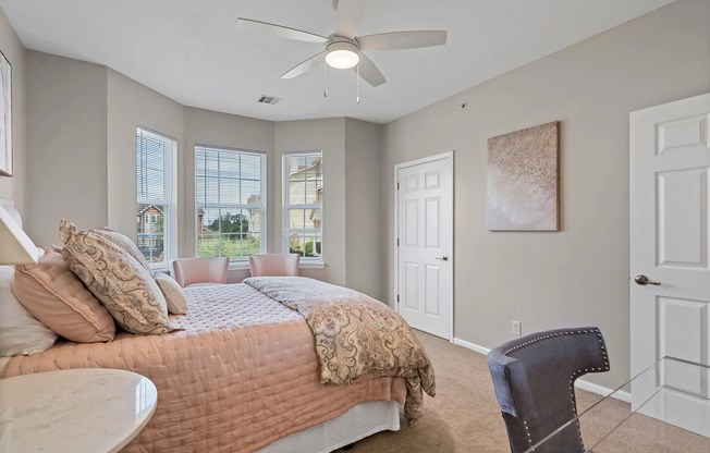Primary bedroom at Kelly’s Ridge Apartments in Columbia, MO, featuring a large window with natural light, neutral-toned walls, a plush bed with layered pillows, stylish bedside tables with lamps, a ceiling fan, and contemporary decor for a serene, inviting atmosphere.