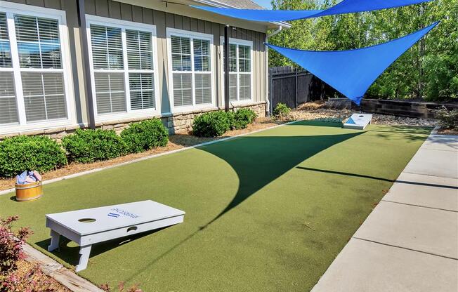 Outdoor grassy area with a cornhole game under triangular shade sails hanging above near a building