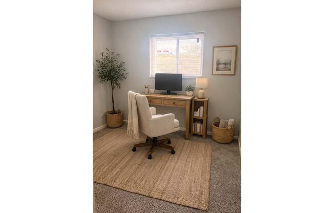 A cozy home office featuring a wooden desk with a computer monitor, a comfortable white chair, and a small lamp. Decorated with a framed picture on the wall, a potted plant in the corner, and a woven basket holding supplies. The space is well-lit with natural light from a window, and has a neutral, inviting color scheme.