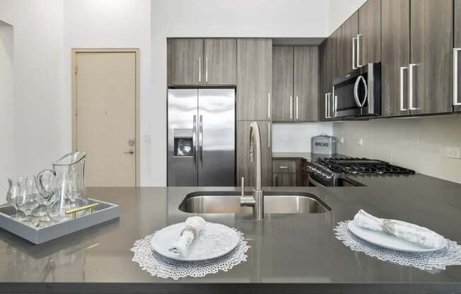 A modern kitchen with a stainless steel refrigerator, a sink, and a countertop with a tray of glasses and two plates.
