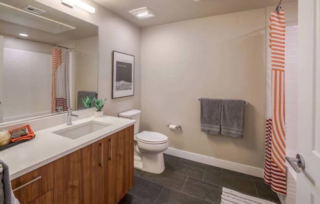 A Bathroom With Luxury Plank Flooring, Large Vanity Mirror, Sink With Wood-Inspired Cabinets, Toilet, And A Shower With Tub. at NoMa Apartments, California, 94596