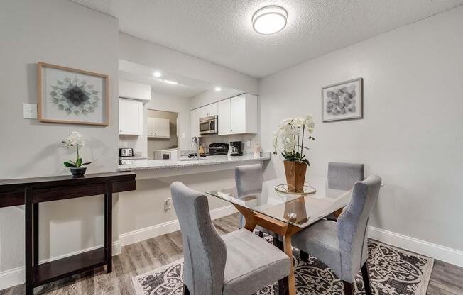 A modern dining area featuring a glass-top table surrounded by four gray upholstered chairs. There is a decorative plant with white flowers on the table, and wall art is visible in the background. The kitchen area is partially visible, showcasing sleek cabinetry and appliances.