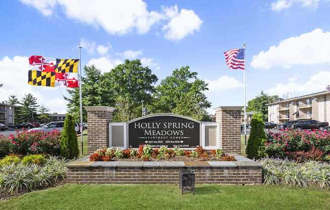 The entrance to Holly Spring Meadows is framed by a brick wall and adorned with a sign, a Maryland state flag, and an American flag.