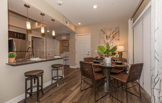 A modern dining area featuring a round table set for four with stylish chairs. In the background, a kitchen bar with pendant lighting, a plant centerpiece, and decorative wall art. The space has warm-toned wooden flooring and soft lighting, creating a cozy and inviting atmosphere.