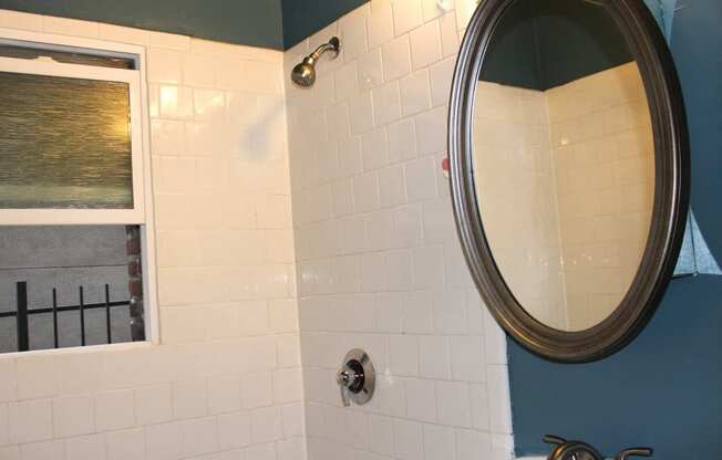 Bathroom in one bedroom brookmore apartment in Pasadena. Shower with white tile and silver fixtures and open window, and oval mirror above pedestal sink