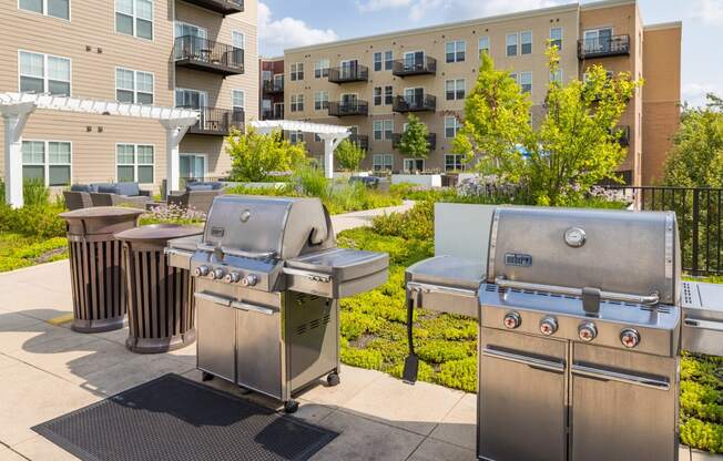 three stainless steel barbecue grills on a patio with apartments in the background