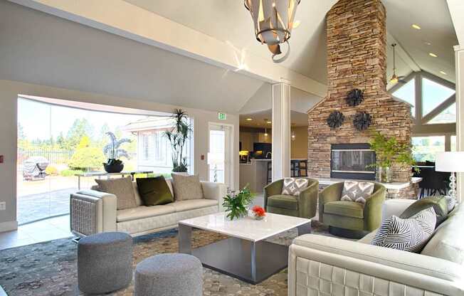 A welcoming resident lounge here at Andorra featuring a vaulted ceiling with modern pendant lighting, a striking stone fireplace, plush sofas and green accent chairs arranged around a sleek coffee table, soft patterned carpeting, decorative plants, and large windows that bring in natural light and views of the outdoor patio.