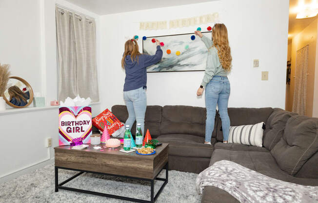 two girls hanging a happy birthday banner on a wall in a living room