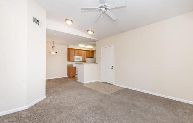 An empty room with beige walls and carpet, featuring a ceiling fan and a small entryway. An open kitchen with wooden cabinets and white appliances is visible in the background. There is a doorway leading to another room and a light fixture hanging from the ceiling. The space is bright and uncluttered.