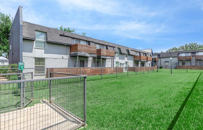 A view of a modern apartment complex with multiple units featuring balconies, surrounded by green lawns and wooden fencing. The sky is clear, and the landscaping appears well-maintained, creating a welcoming outdoor space for residents.
