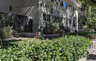 Sidewalk view of stone wall garden beds and mature shade trees in front of Los Robles Apartments in Pasadena, California.