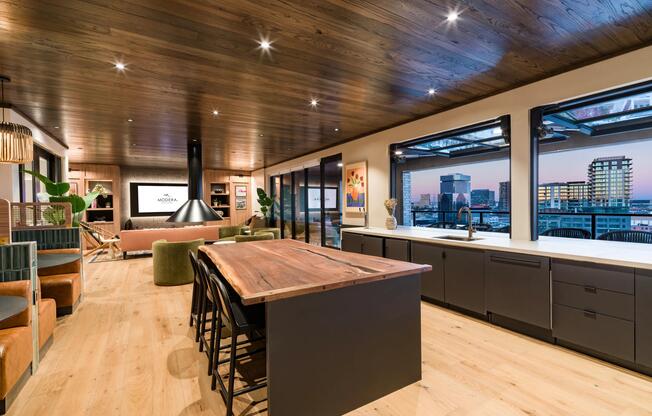 An inviting clubroom kitchen in our sky lounge with a warm wood island, open seating, and expansive windows framing downtown Dallas views.
