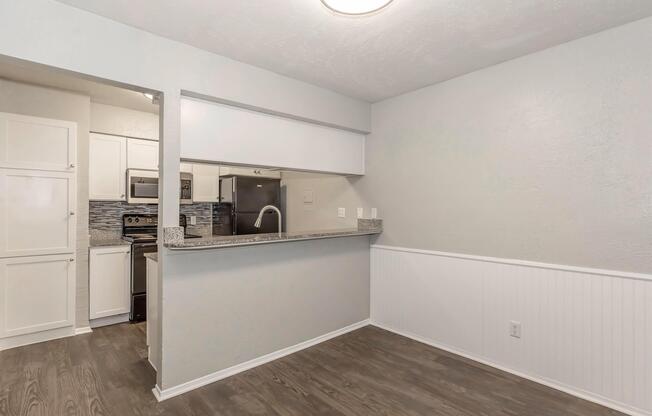 A modern kitchen with a partial wall separating it from a dining area. The kitchen features granite countertops, stainless steel appliances, and light-colored cabinetry. The dining area has wood-like flooring and a light fixture overhead, creating a bright, inviting space.