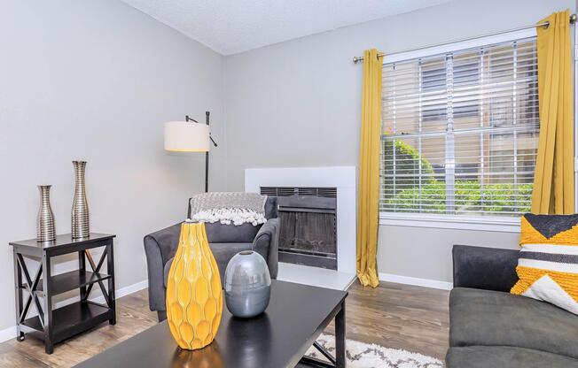 A cozy living room featuring a black armchair and a dark sofa, with a coffee table holding a decorative gray and yellow vase. A modern floor lamp stands beside the chair, and a window with yellow curtains offers natural light. The walls are painted light gray, creating a warm and inviting atmosphere.