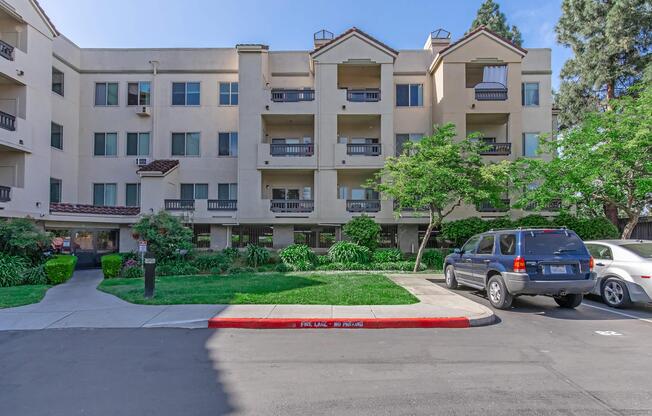 A multi-story residential building with a light beige exterior, featuring balconies on the upper floors. The landscaped area includes trees and shrubs. A parking lot is visible in the foreground with a silver SUV parked, and a red curb marked "FIRE LANE – NO PARKING." Clear blue sky in the background.