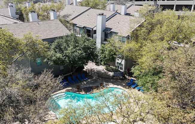 A swimming pool surrounded by lush trees and apartment buildings at The Park on Preston Apartments in Dallas, TX
