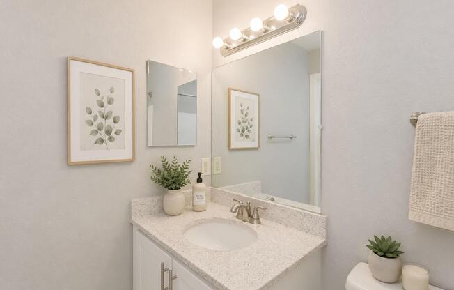 A modern bathroom with light gray walls, featuring a white vanity with a sink, a large mirror, and decorative wall art. There is a plant on the counter and a towel hanging nearby. Soft lighting is provided by a fixture above the mirror, creating a bright and inviting atmosphere.
