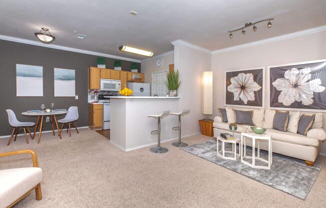 A modern, cozy apartment with an open layout. The living area features a beige sofa, decorative tables, and a rug, while the dining area includes a small table and chairs. The kitchen is equipped with wooden cabinets and white appliances. Two floral artworks adorn the walls, contributing to the stylish decor.