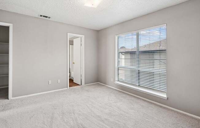 A room with a white carpet, a window with blinds, and a door leading to another room.