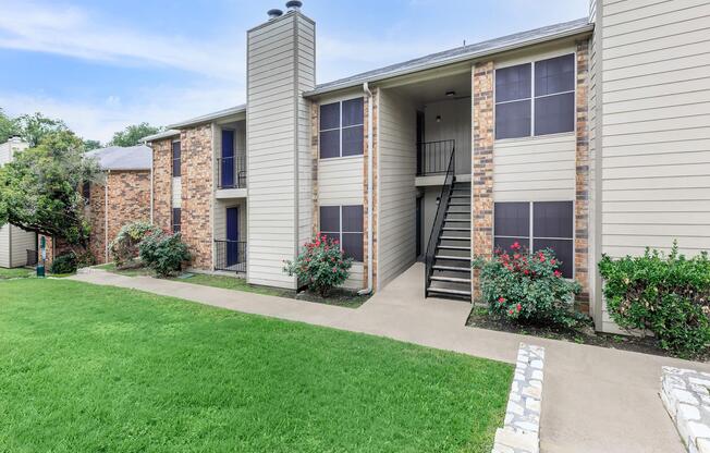 A view of an apartment community featuring two-story buildings with light-colored siding and brick accents. The pathway is lined with well-maintained grass and flower bushes, leading to stairs that provide access to the upper level of the building. The sky is bright and clear.