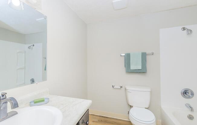 A clean and modern bathroom featuring a white sink with a light-colored countertop, a toilet, and a bathtub with a shower. The walls are painted light gray, and there is a mirror above the sink. A green towel and washcloth hang on a bar next to the sink, and a small green soap dish is on the countertop.
