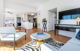 A modern, bright living space featuring a comfortable white chair, a stylish marble coffee table with a patterned rug, and sleek furniture. The open layout includes a kitchen area in the background, with natural light streaming in from large windows, creating an inviting atmosphere.