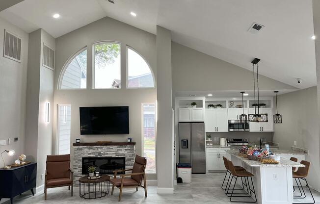 A modern open-concept living space featuring high ceilings, a large window, and stylish decor. There is a cozy seating area with two brown chairs and a coffee table, a sleek entertainment unit, and a kitchen with white cabinetry, stainless steel appliances, and a dining area with bar stools. Light floods the room, creating an inviting atmosphere.