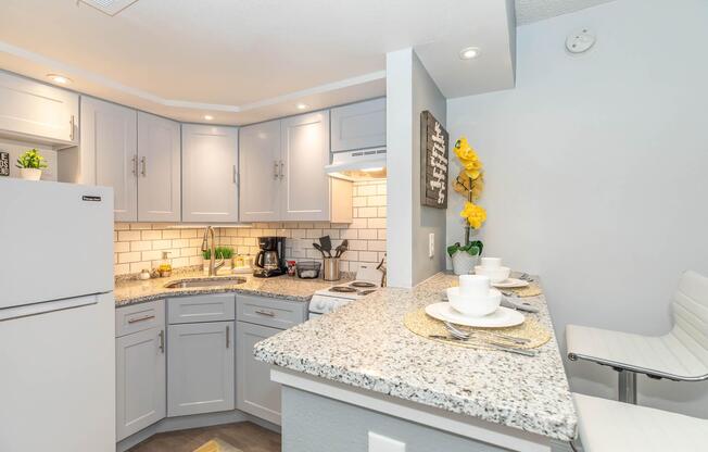 Modern kitchen with light gray cabinets and white tile backsplash. Features a granite countertop with breakfast bar seating for two. Appliances include a white refrigerator and stove. The space is decorated with plants and yellow flowers, creating a welcoming atmosphere. Soft lighting enhances the cozy feel.