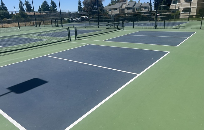 A view of several tennis courts with blue and green surfaces under a clear blue sky. The courts are empty, with surrounding fencing and some nearby residential buildings visible in the background.