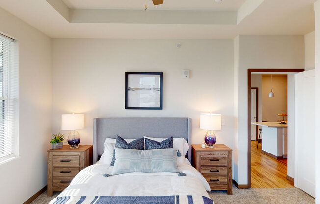 A comfortable bedroom here at Van Buren East with soft carpeting and neutral tones. A large window brings in natural light, while a cozy bed and bedside lighting create a warm, inviting atmosphere.