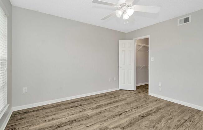 A light gray room with a ceiling fan, featuring a closet with open doors. The walls are unadorned, and large windows let in natural light, highlighting the brown wooden-style flooring. The space is empty, providing a blank canvas for decoration.