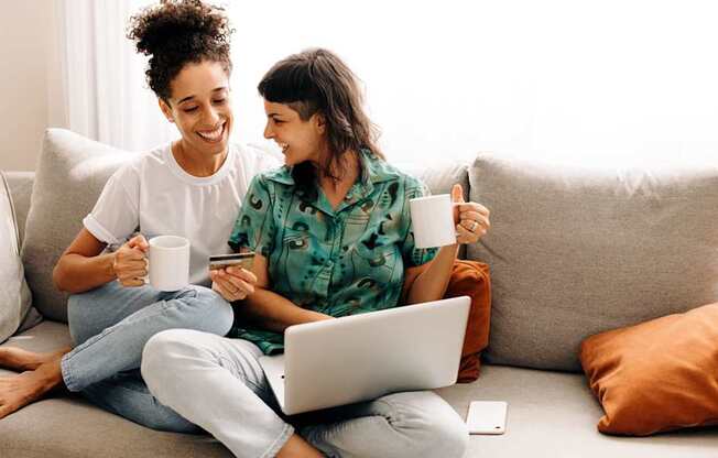 two women sitting on a couch drinking coffee while using a laptop computer at North Grove, Riverside, CA  