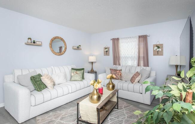 A bright and cozy living room featuring two white sofas adorned with decorative pillows. A wooden coffee table holds vases with flowers, and a soft area rug is beneath. Wall decorations include framed floral art and shelves with small plants. Natural light filters through a window covered with sheer curtains.