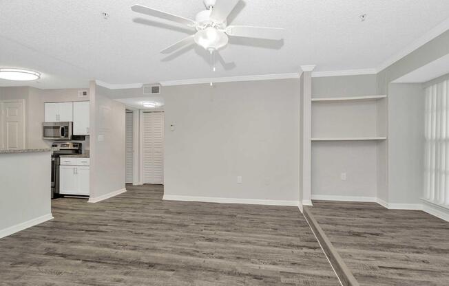 Spacious and bright living area with light-colored walls, ceiling fan, and modern laminate flooring. An open kitchen with stainless steel appliances and a bar area is visible. A built-in shelf is placed along one wall, and there are large windows allowing natural light to fill the space.
