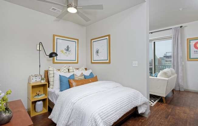 Furnished bedroom with white sheets, nightstand and ceiling fan, opens to living room