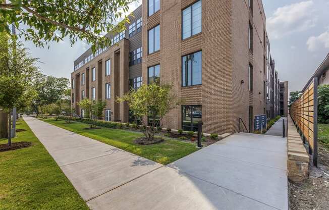 A long concrete walkway leads to a brick building with a green lawn on the side.