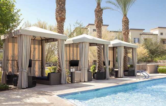 Four palm trees are behind a pool with four sun shelters.