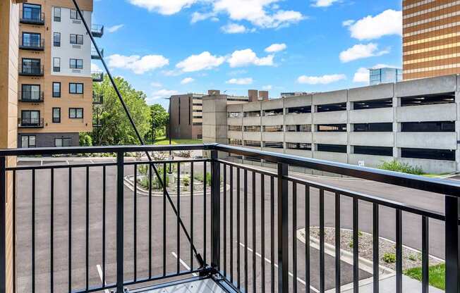 A balcony with a black railing overlooks a parking garage and buildings.