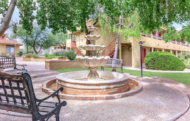 A tranquil courtyard featuring a tiered stone fountain surrounded by lush greenery. Nearby, there are benches for seating and staircases leading to upper-level buildings. The setting has a peaceful ambiance, ideal for relaxation or socializing.