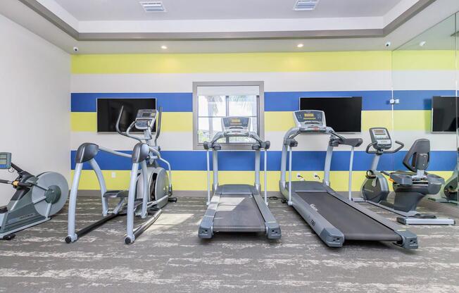 A modern gym interior featuring three exercise machines: two treadmills and an elliptical. The walls are painted in alternating yellow and blue stripes, and there are two flat-screen TVs mounted on the wall. Large windows provide natural light, and the flooring is a plush, neutral-toned carpet.