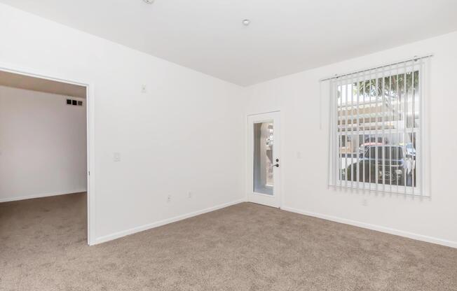 A spacious, empty room with beige carpet, white walls, and a doorway leading to an exterior entrance. A window with blinds allows natural light to enter. The adjoining room is visible through an open doorway, suggesting a layout that connects two areas.