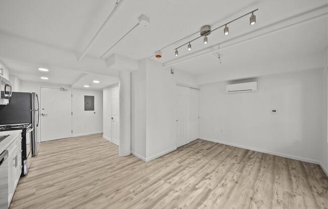 A modern, empty apartment interior featuring a kitchen on the left with stainless steel appliances and a living area on the right. The space has light-colored wooden flooring, white walls, and recessed lighting, creating a bright and airy atmosphere. A door leads to the hallway, and there's an air conditioning unit on the wall.