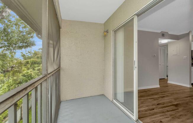 A small balcony with a sliding glass door open to an indoor space. The balcony features a light-colored floor and walls, with a view of greenery outside. The interior room is visible through the door, showcasing a light-colored wall and wooden flooring.