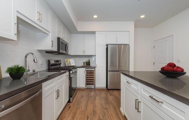 Modern kitchen featuring white cabinetry, stainless steel appliances, a sleek dark countertop, and hardwood flooring. There is a wine cooler and a bowl of red apples on the counter, creating a clean and inviting space. Bright, natural light enhances the contemporary design.