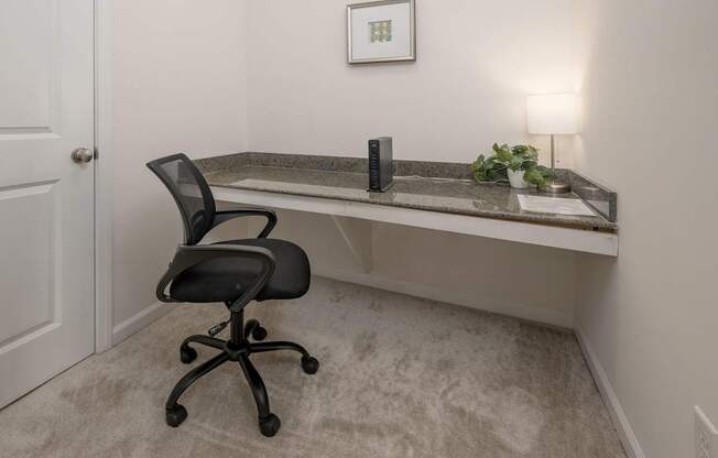 A black office chair is in front of a desk with a plant and a picture on the wall.