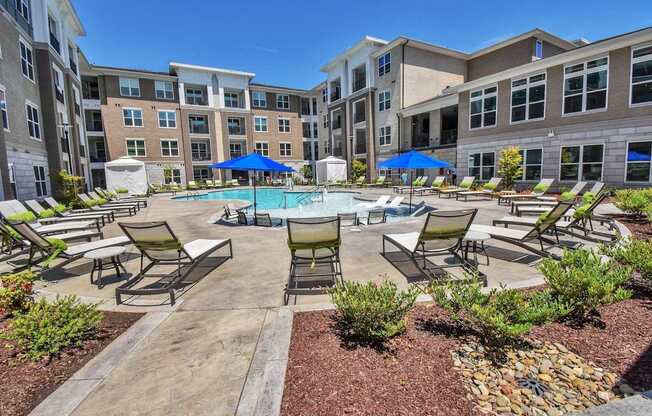 A pool surrounded by chairs and umbrellas in front of apartment buildings.