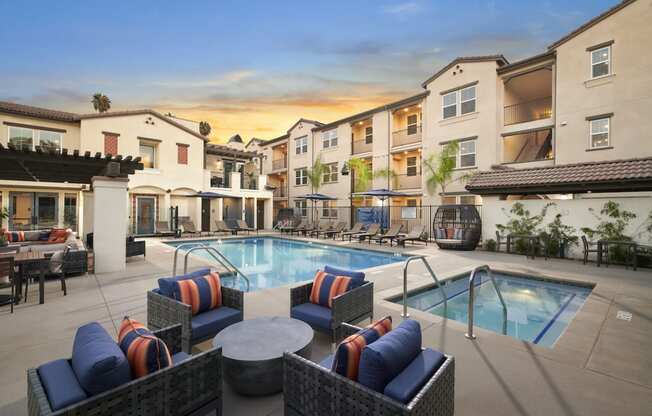 Apartments in Riverside, CA for Rent - Lincoln Village - Large Pool and Hot Tub, with Lounge Chairs, Outdoor Seating Area with Coffee Table, Covered Sofas, and Umbrellas, with White Building Exterior in the Background